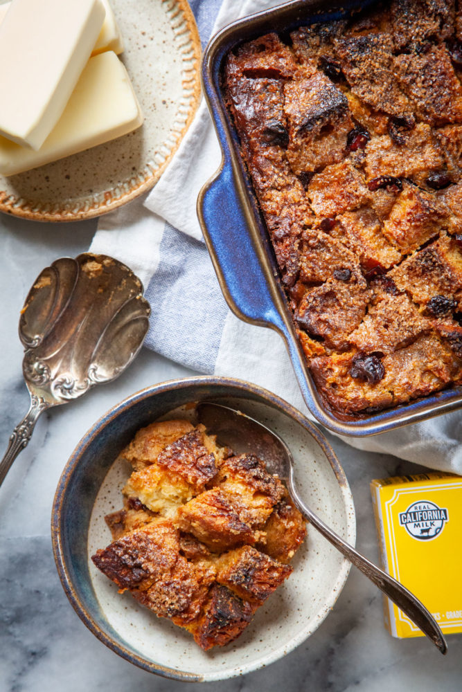 A bowl of pumpkin bread pudding with a pan of more bread pudding and a plate of butter sticks near it.