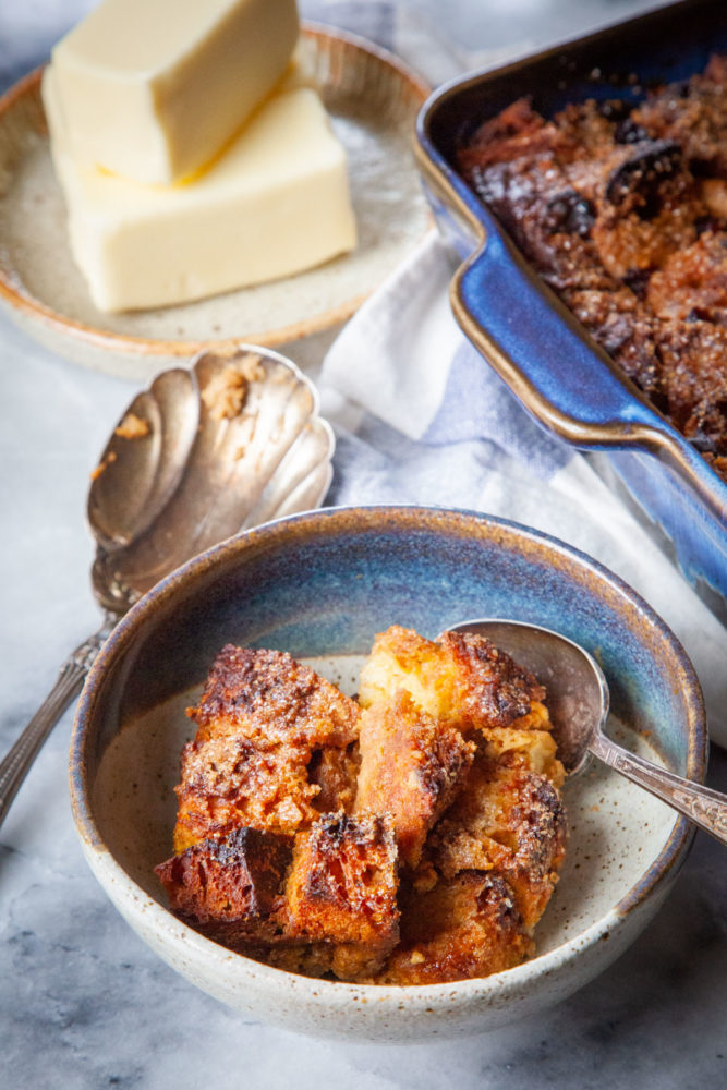 A bowl of pumpkin bread pudding with a pan of more bread pudding and a plate of butter sticks near it.