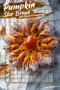 A pumpkin star bread with dried cranberries filling sitting on a gold wire cooling rack with a kitchen towel underneath.