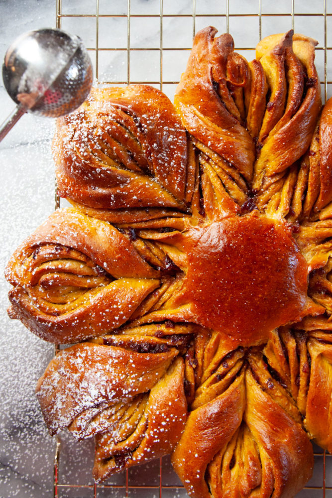 A pumpkin star bread with dried cranberries filling sitting on a gold wire cooling rack being dusted with powdered sugar.