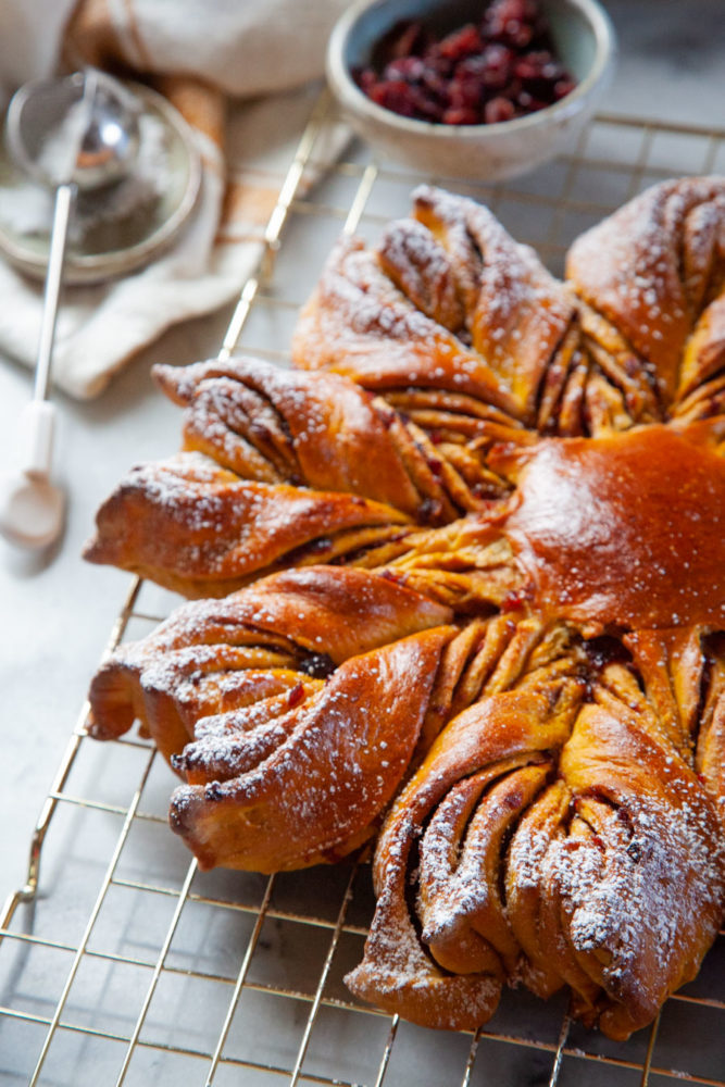 A pumpkin star bread with dried cranberries filling sitting on a gold wire cooling rack with dried cranberries and a powdered sugar wand behind it.