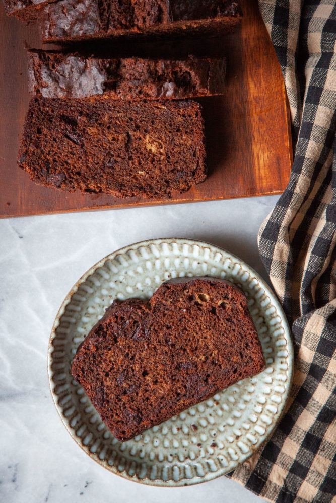 A slice of chocolate banana bread on a plate with more banana bread on a cutting board next to it.