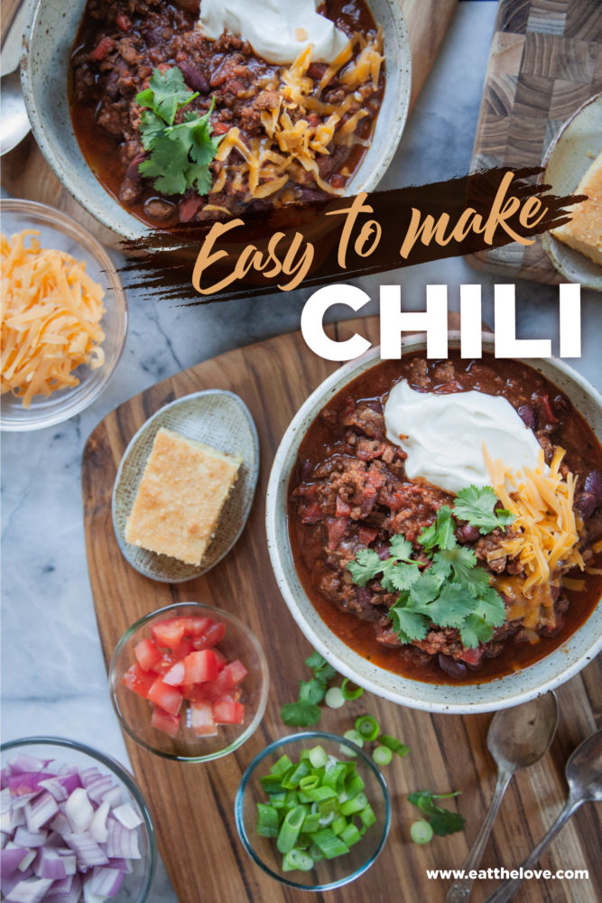 Two bowls of chili surrounded by toppings like chopped tomatoes, shredded cheese and red onions.