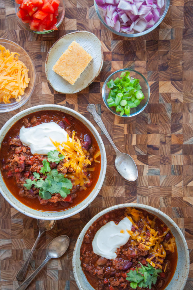 Two bowls of chili surrounded by toppings like chopped tomatoes, shredded cheese and red onions.