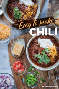 Two bowls of chili surrounded by toppings like chopped tomatoes, shredded cheese and red onions.