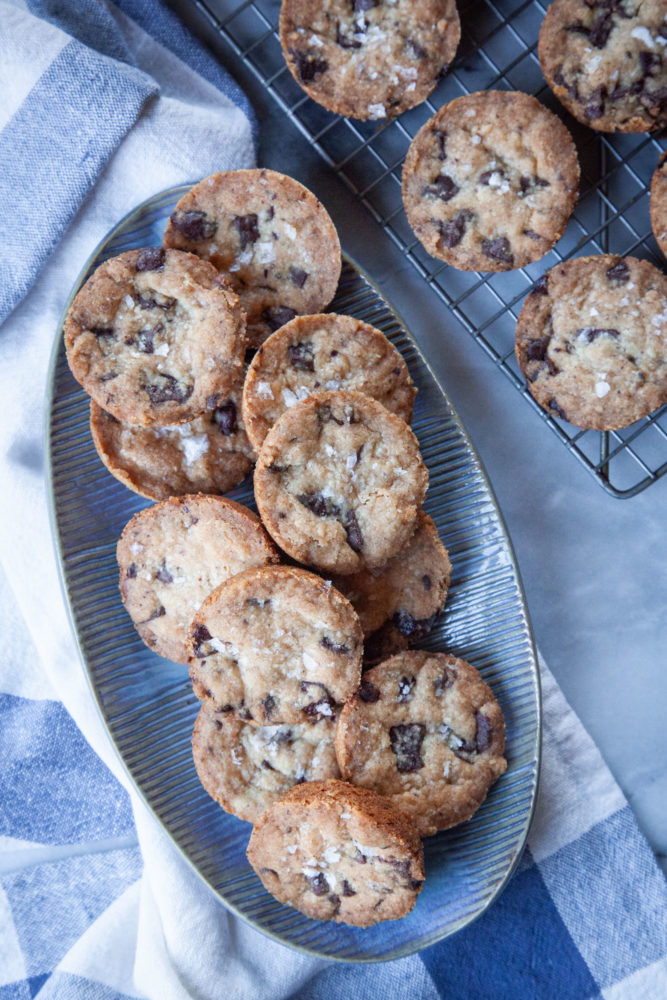 Pile of shortbread chocolate chip cookies on a blue oval plate with more cookies on a wire cooling rack next to it.