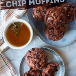 Two double chocolate chip muffins on a serving plate with a cup of tea a platter of more muffins next to it.