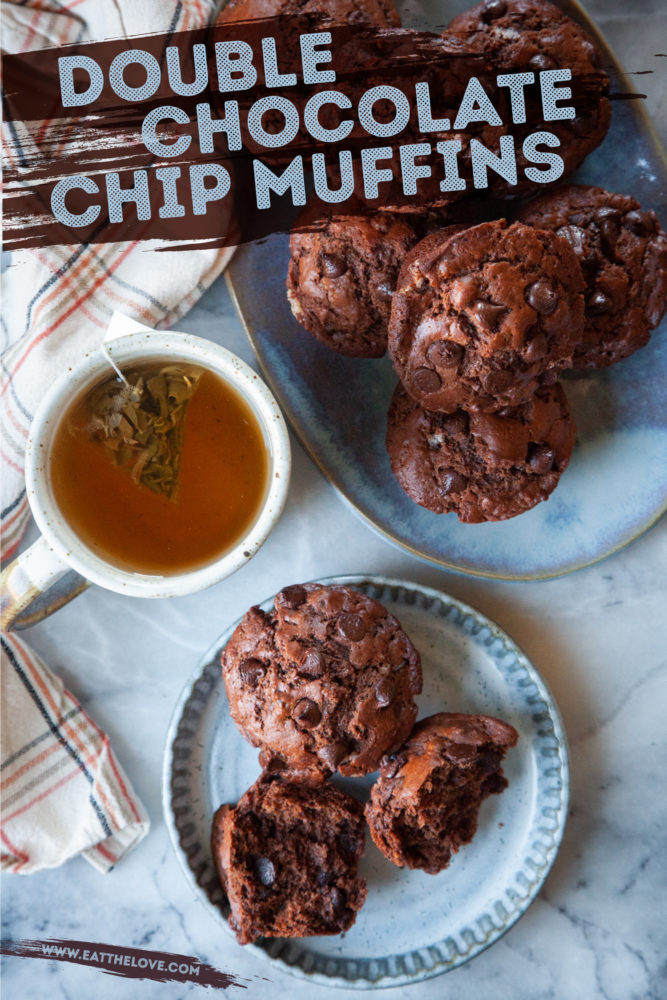 Two double chocolate chip muffins on a serving plate with a cup of tea a platter of more muffins next to it.