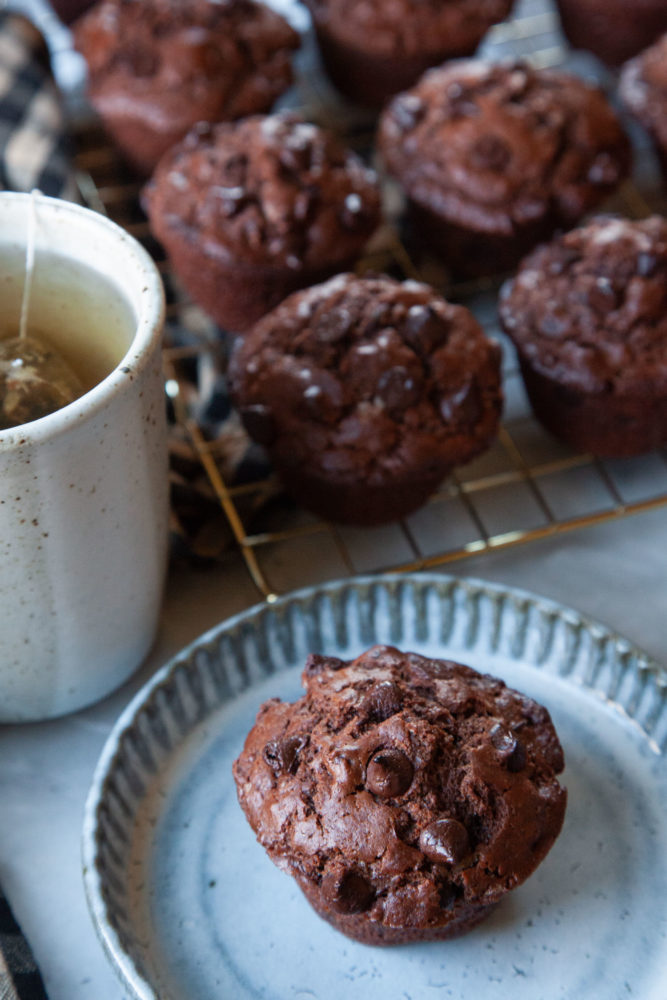 A double chocolate chip muffin on a plate, with a mug of tea and a wire cooling rack of more muffins behind it.