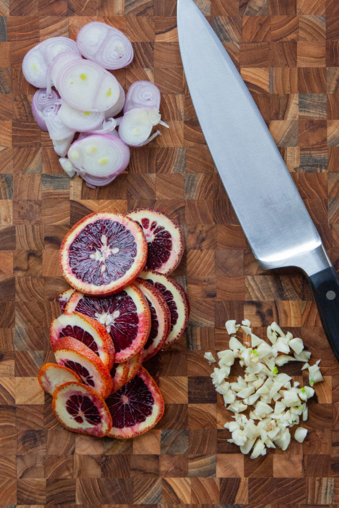 Sliced shallots, blood oranges and chopped garlic on a cutting board with a chef knife next to the ingredients.