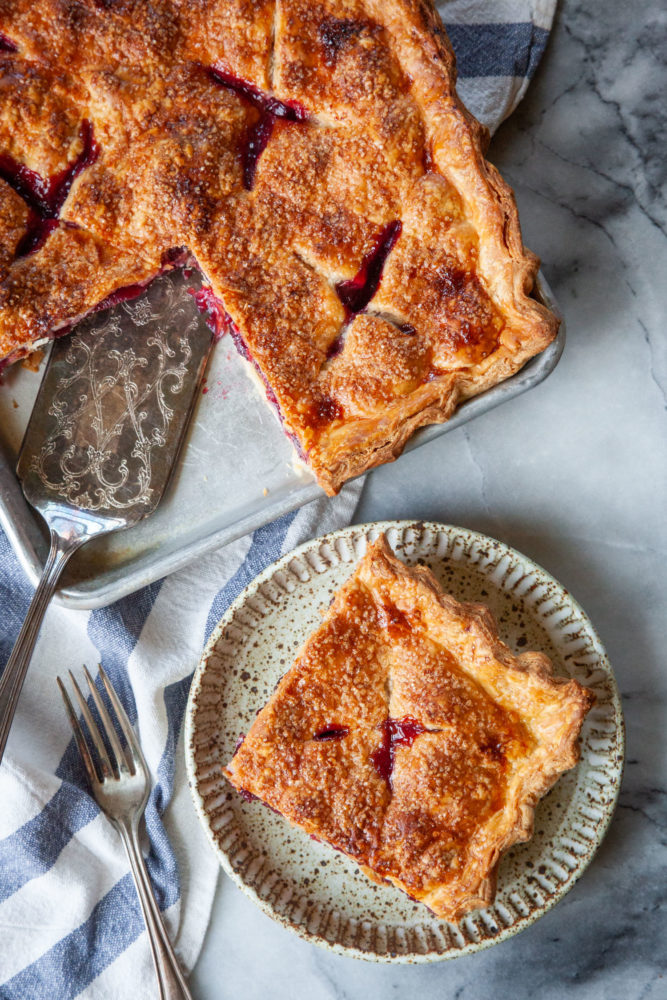 A slice of cherry slab pie on a plate, with the remaining slab pie in a pan next to it.