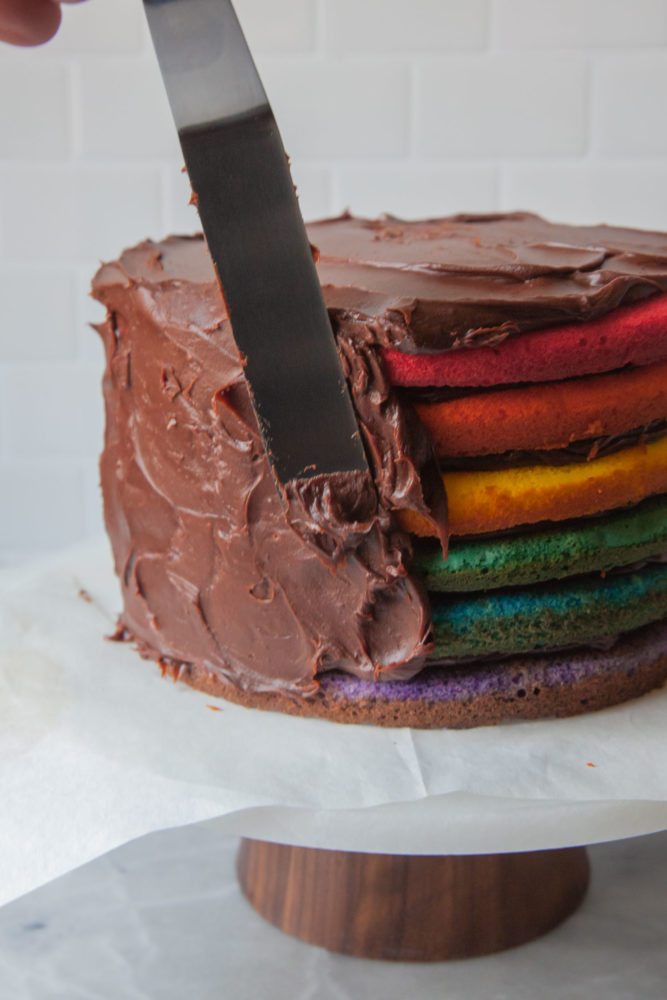 A hand frosting a rainbow layer cake with chocolate frosting on a cake stand.