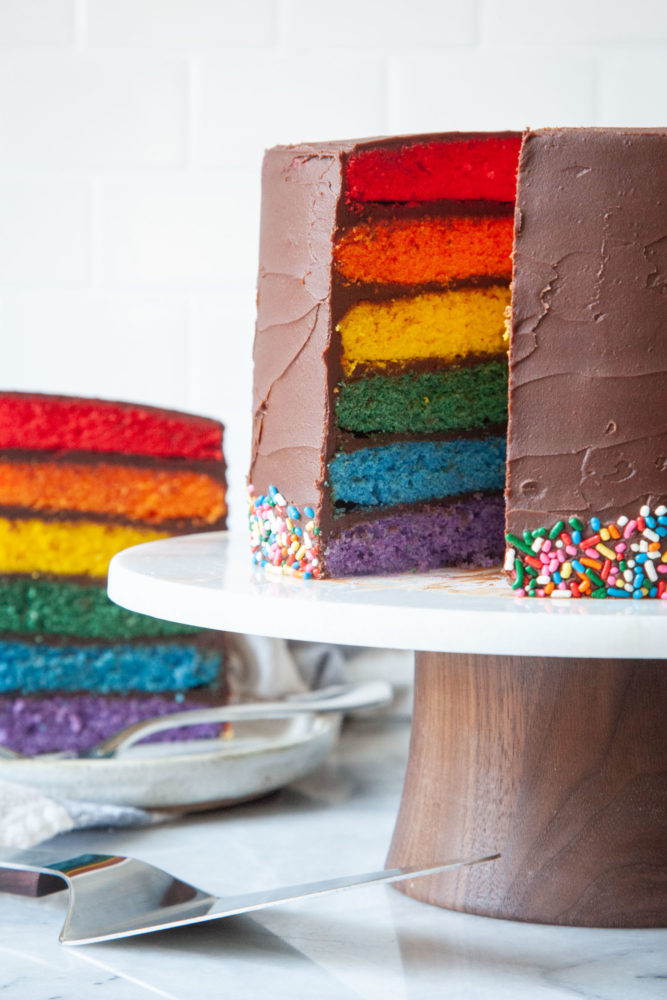 A rainbow layer cake on a cake stand with a slice cut out, and the slice on a plate behind it.