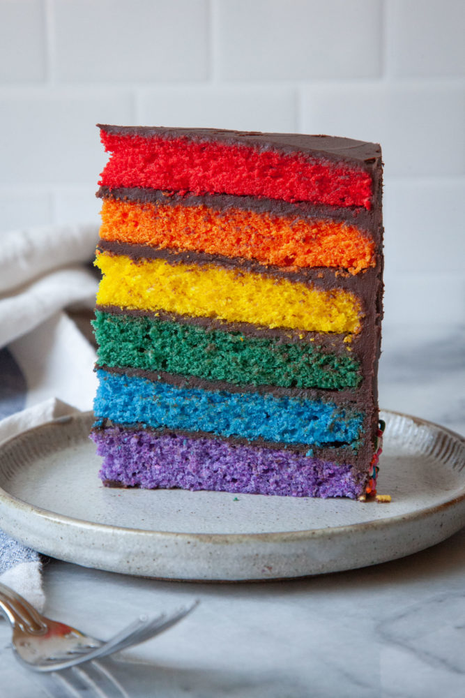 A slice of rainbow layer cake with chocolate frosting on a plate.