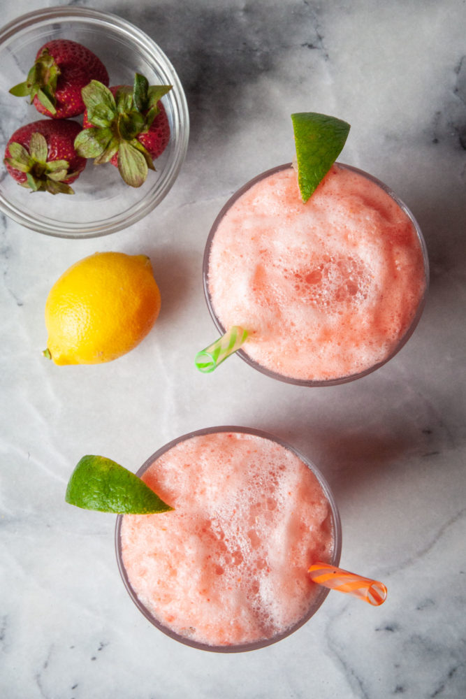 A top down view of two glasses of sparking strawberry lemonade with a bowl of bowl of strawberries and a lemon next to them.