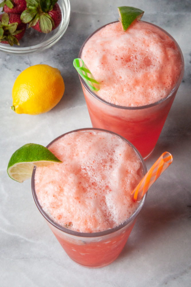 Two glasses of sparking strawberry lemonade with straws sticking out of them. A lemon and a bowl of strawberries are behind them.