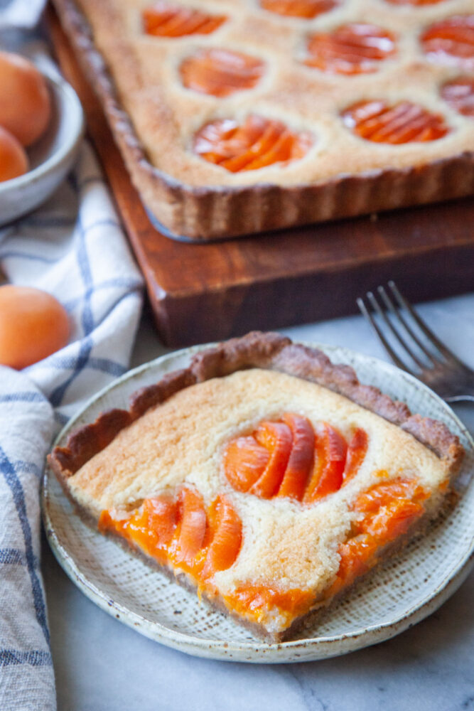 A slice of apricot frangipane on a plate, with the remaining tart behind it.