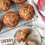 A cherry almond muffin split in half on a plate, with more muffins on a wire rack next to it.