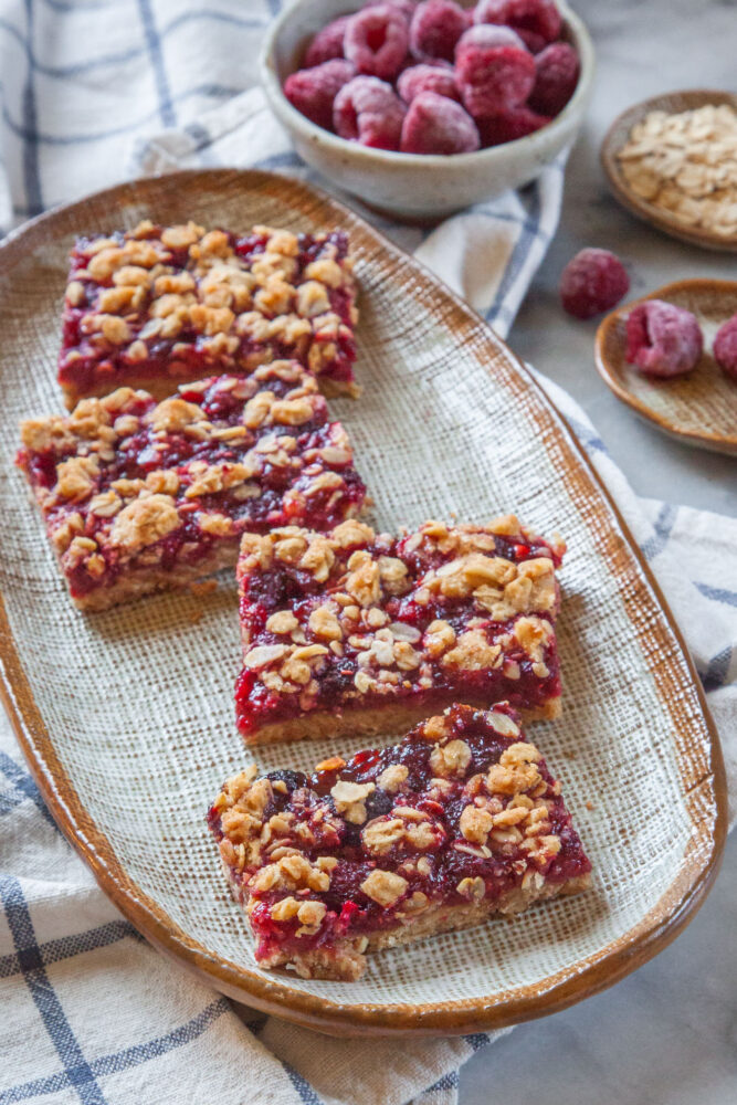Raspberry oat bars on an oval plate, with a small bowl of frozen raspberries and a small plate of oats behind it. 