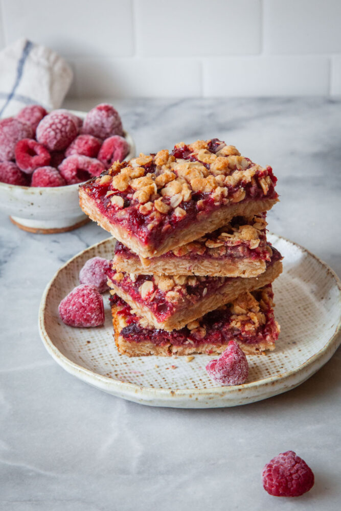 A stack of raspberry oat bars on a small plate, with a couple of frozen raspberries next to it, and a small bowl or frozen raspberries behind it.