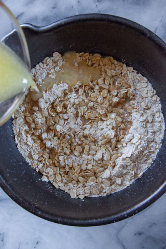 A glass measuring cup drizzling melted butter into a mixing bowl with oat crust ingredients in it.