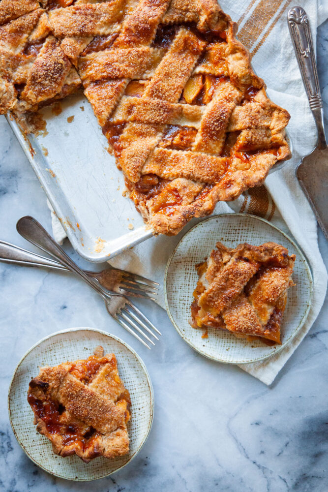 Two slices of peach slab pie on plates, with the rest of the pie next to it. 