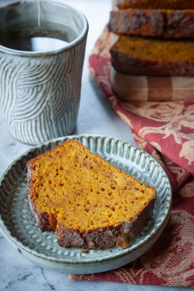 A slice of pumpkin bread on a plate, with a cup of tea behind it and the remaining pumpkin bread on a cutting board next to the tea.
