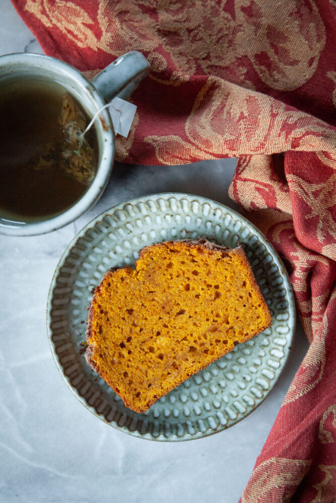 A slice of pumpkin bread on a plate, with a cup of tea next to it.