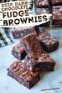 A stack of fudge brownies on a marble counter, with more brownies on a wire cooling rack behind them.