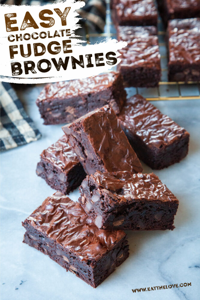 A stack of fudge brownies on a marble counter, with more brownies on a wire cooling rack behind them.