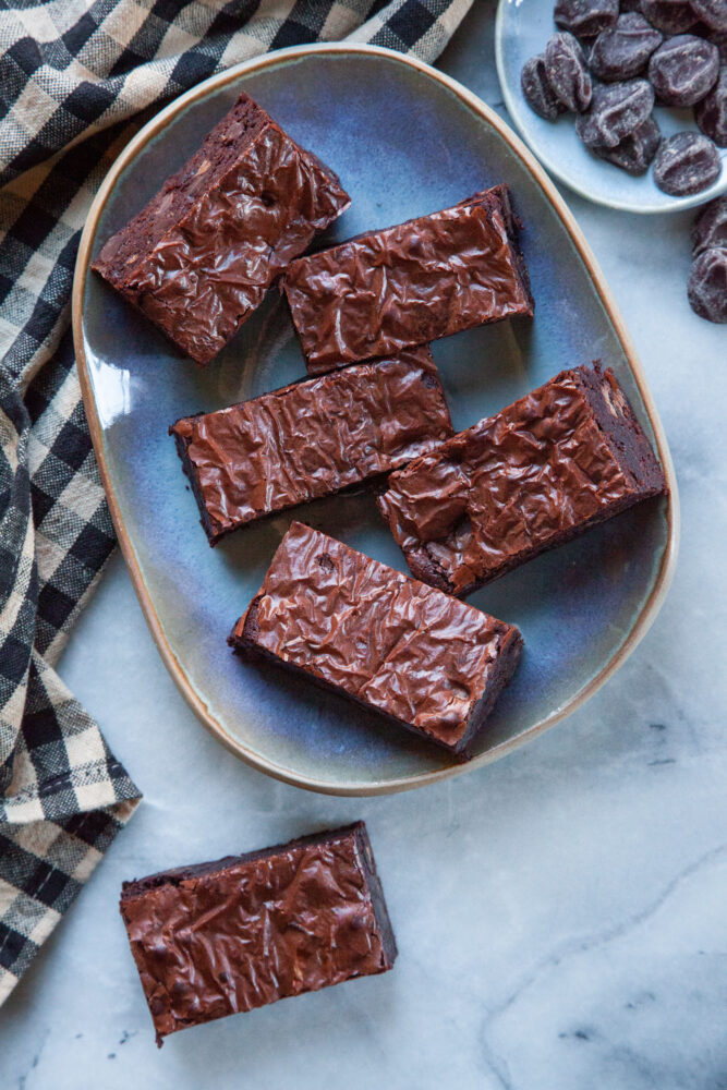 Fudge brownies on a plate, with a brownie on the marble countertop next to it. There is a small plate of chocolate chips next to them.