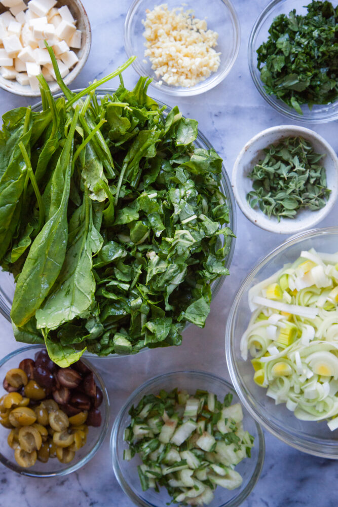 Ingredients for the green shakshuka base in individual bowls, including chopped leeks, chopped garlic, leafy greens, feta cheese, herbs, olives.