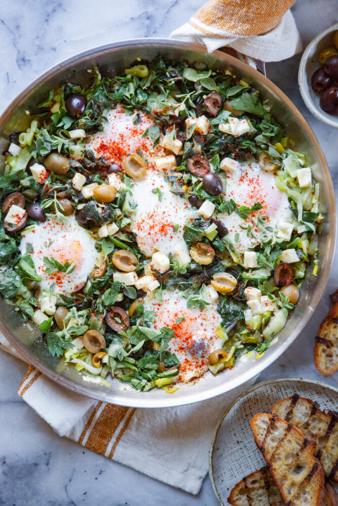 A pan of green Shakshuka with a plate of toast and a bowl of ripe olives next to it.