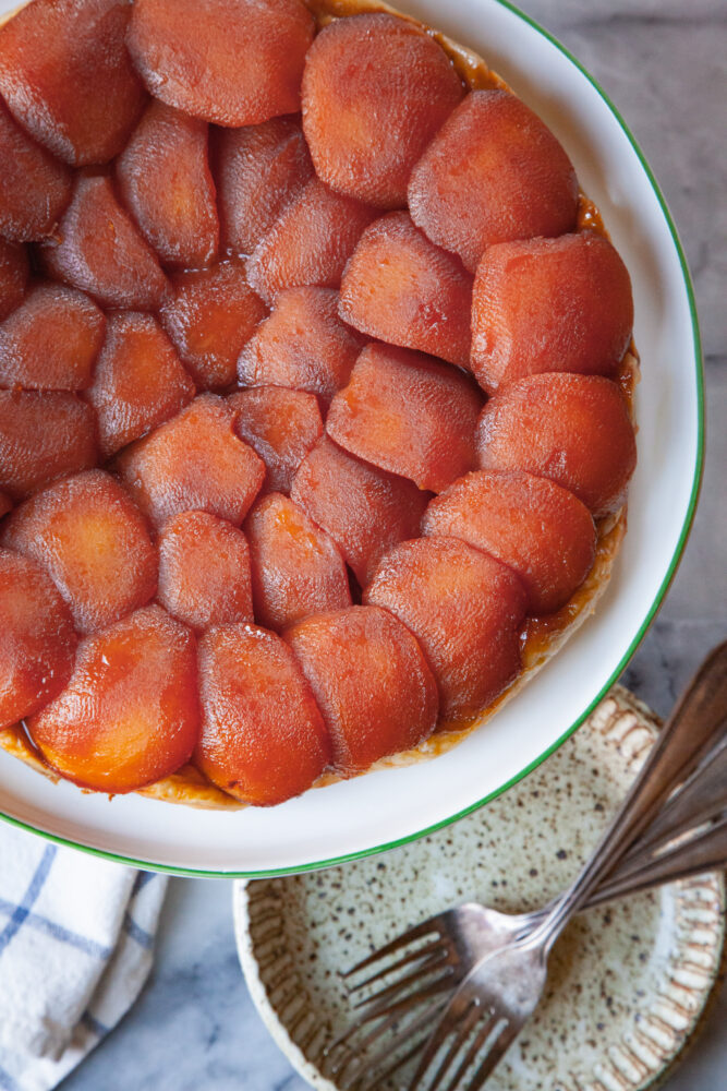 A whole tarte tatin on a cake stand, with a dessert plate and forks under it.