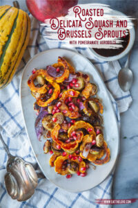 A plate with roasted Delicata squash and Brussels sprouts with pomegranate agrodolce drizzled over it. A pomegranate and delicate squash are sitting next to the plate.