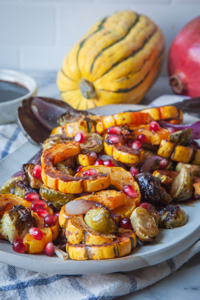 A plate of roasted Delicata squash and Brussels sprouts with pomegranate agrodolce drizzled over it. Behind it is a Delicata squash and a pomegranate.