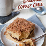A piece of cinnamon streusel coffee cake on a small plate, with a mug of tea behind it.