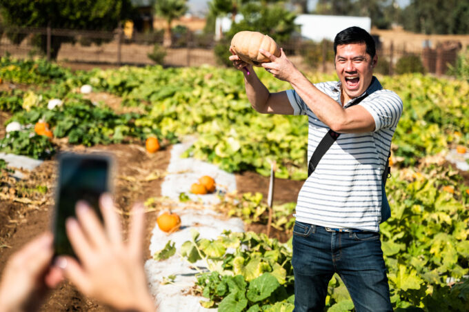 A man standing in a pumpkin field holding up a pumpkin that he picked off a vine. A hand is taking a photo of him with their mobile phone.