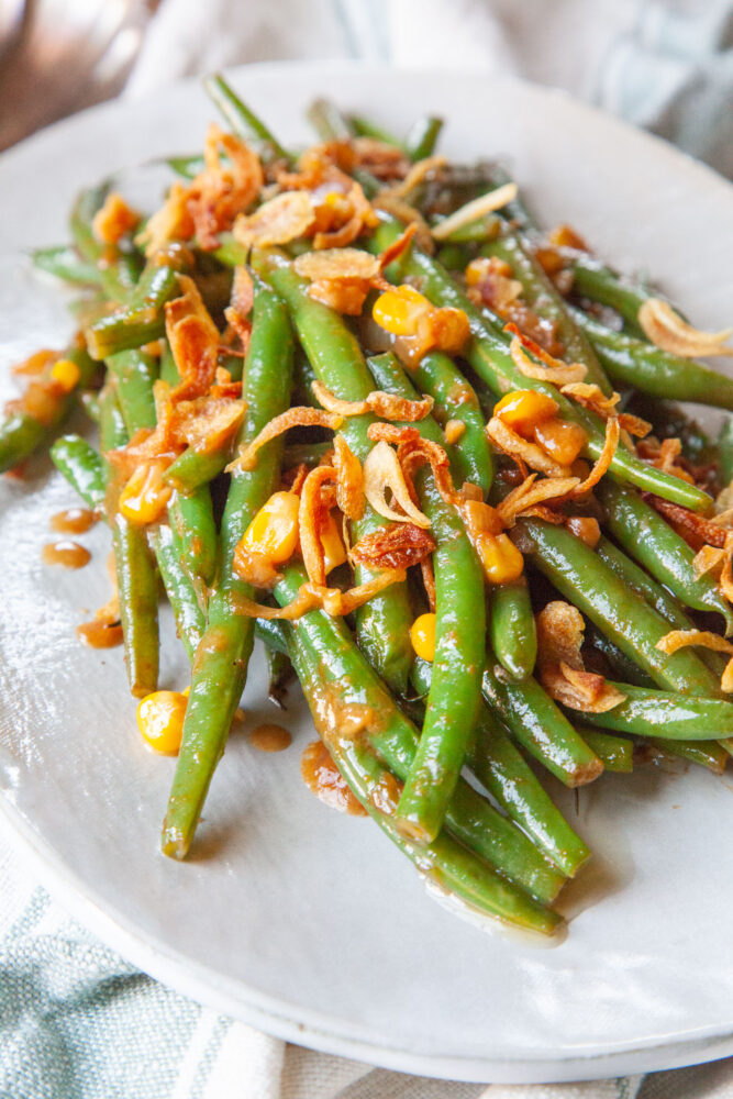 An oval plate with miso butter glazed green beans with corn and fried shallots on it.