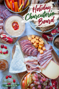 A holiday charcuterie board with cheese, charcuterie, hummus, red bell peppers, carrots, apples and other fruit, as well as drinks on a table.