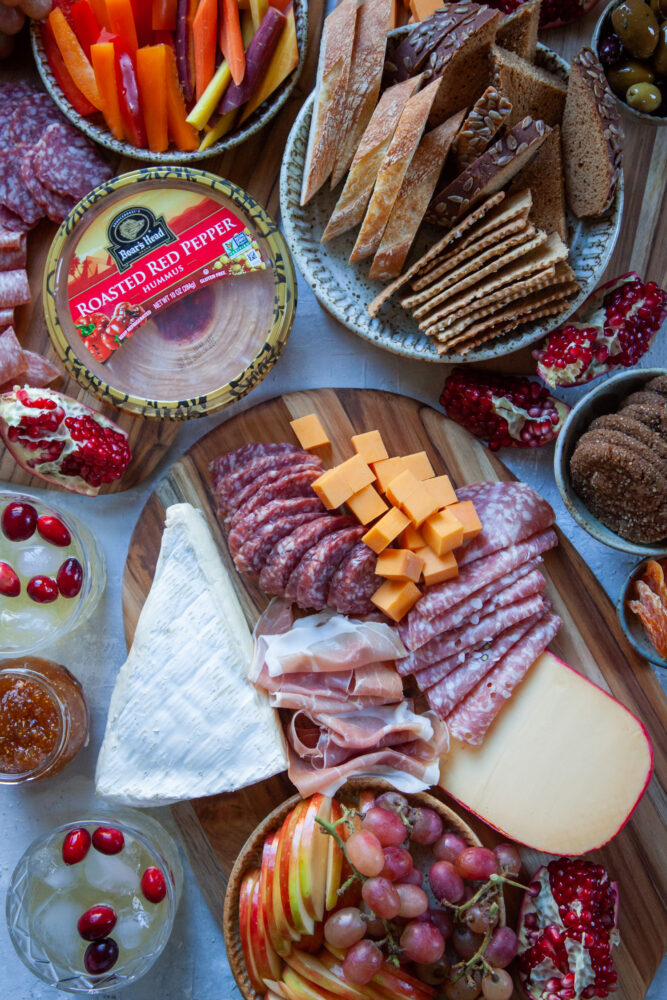 A holiday charcuterie board with cheese, charcuterie, hummus, red bell peppers, carrots, apples and other fruit, as well as drinks on a table.