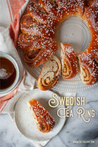 A slice of Swedish tea ring coffee cake on a white plate, with the remaining tea cake on a plate next to it. There is a cup of tea next to the cake.