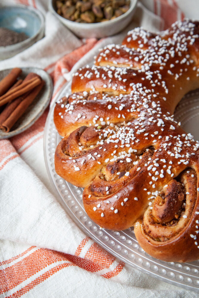 A Swedish tea ring on a glass plate, with an orange and white striped cloth napkin under it. Next to the tea ring are small bowls and plates holding cinnamon sticks, ground cardamom and pistachios, all ingredients used in the filling of the tea ring.