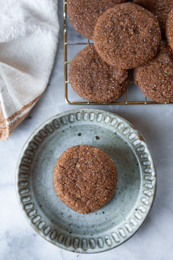 A single chewy ginger molasses cookie on a small plate, with more ginger molasses cookies on a wire cooling rack near it. 