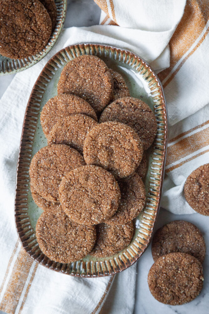 Chewy ginger molasses cookies on an oval plate onto of a cloth napkin, with more cookies next to it.