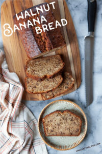 A slice of walnut banana bread on a ceramic plate. Next to the plate is the remaining banana bread loaf on a wooden board, with a serrated knife next to it. There is a cloth napkin next to the banana bread.