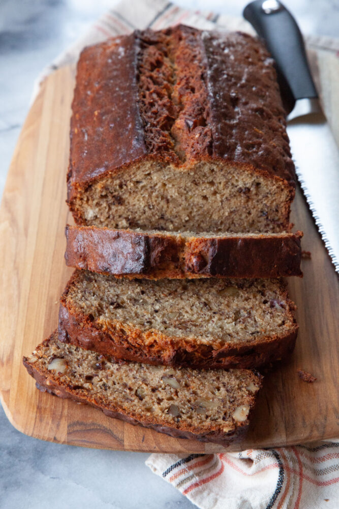 A loaf of sliced walnut banana bread on a wooden serving board with a serrated knife next to it.