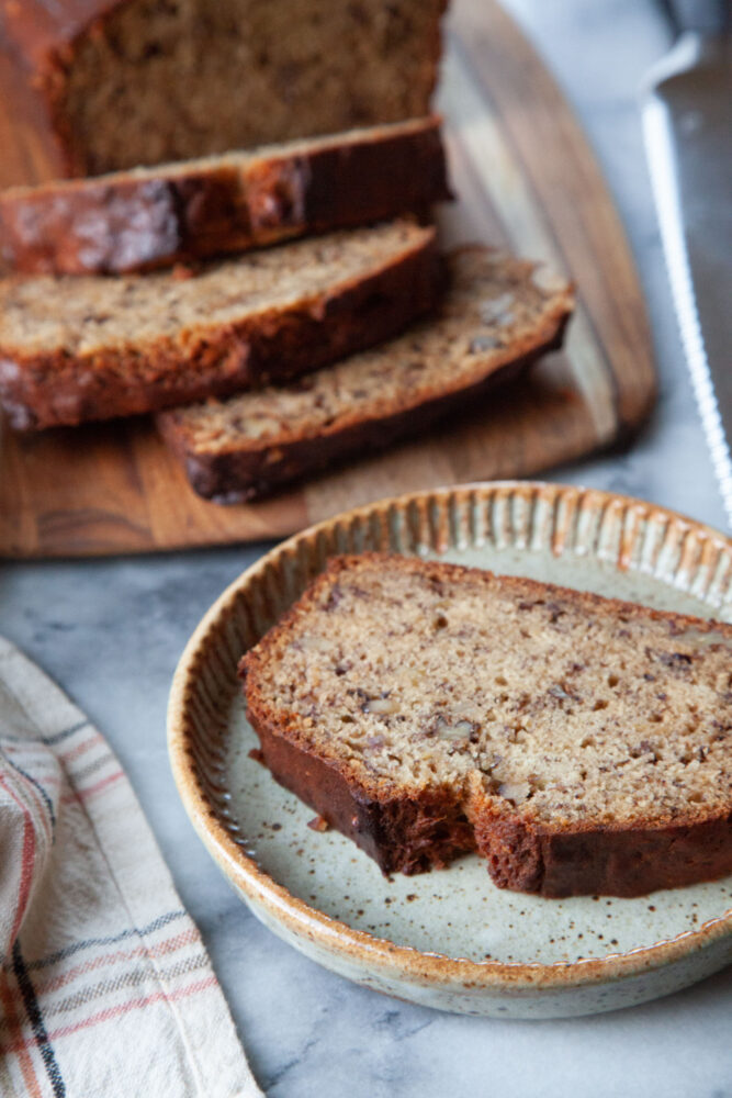 A slice of walnut banana bread on a small ceramic plate, with the remaining banana bread on a wooden serving board behind it.