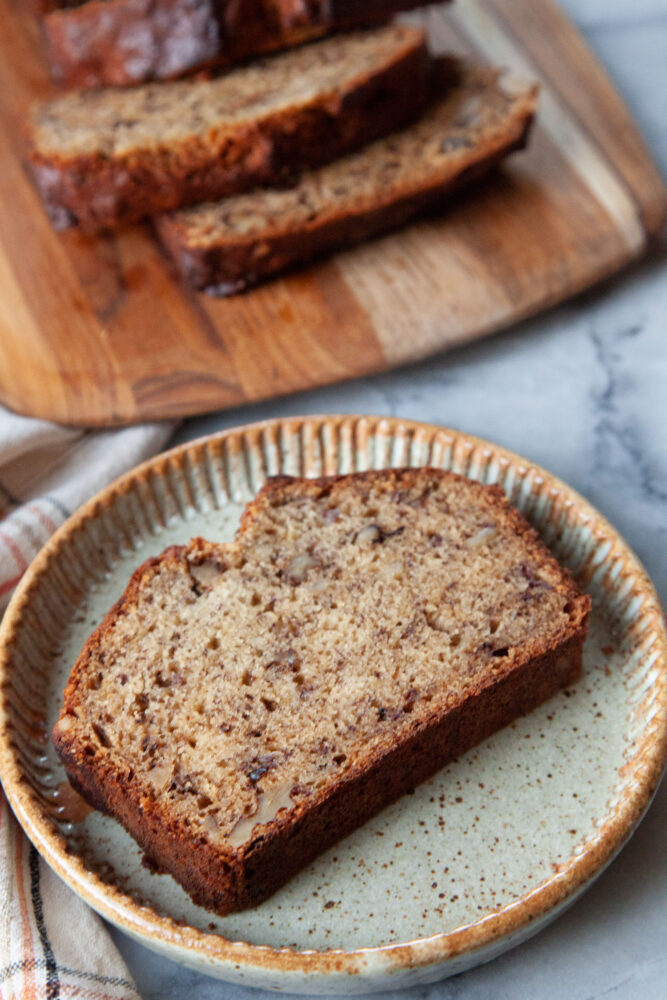 A slice of walnut banana bread on a plate, with the remaining loaf of bread on a wooden serving board behind it.