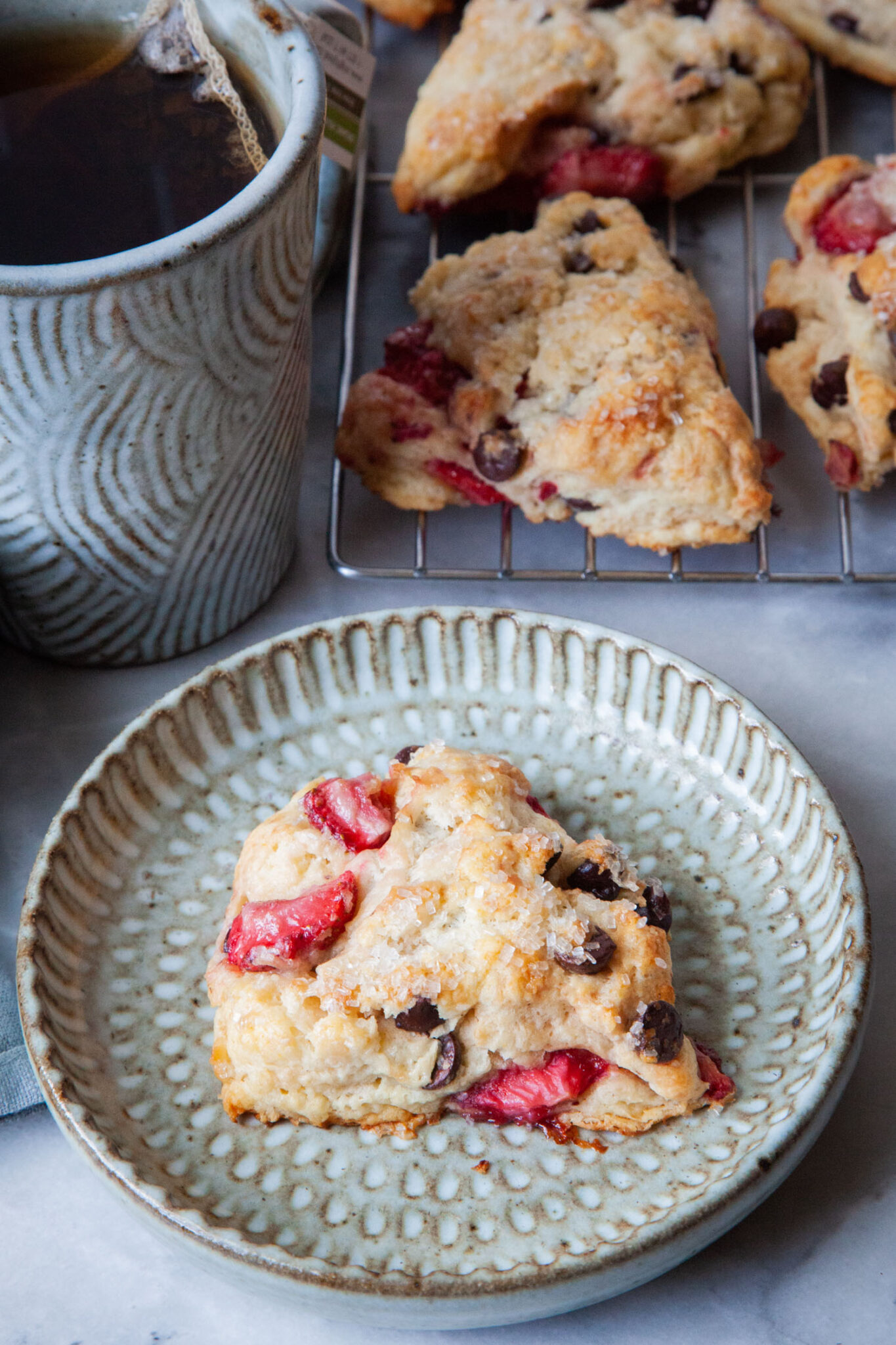  A chocolate and strawberry scone on a plate, with a mug of tea and a wire rack of more scones behind it.